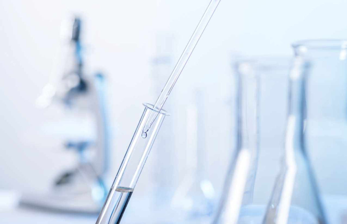 Laboranalyse einer Wasserprobe – Pipette über Reagenzglas, Mikroskop und Glaskolben im Hintergrund