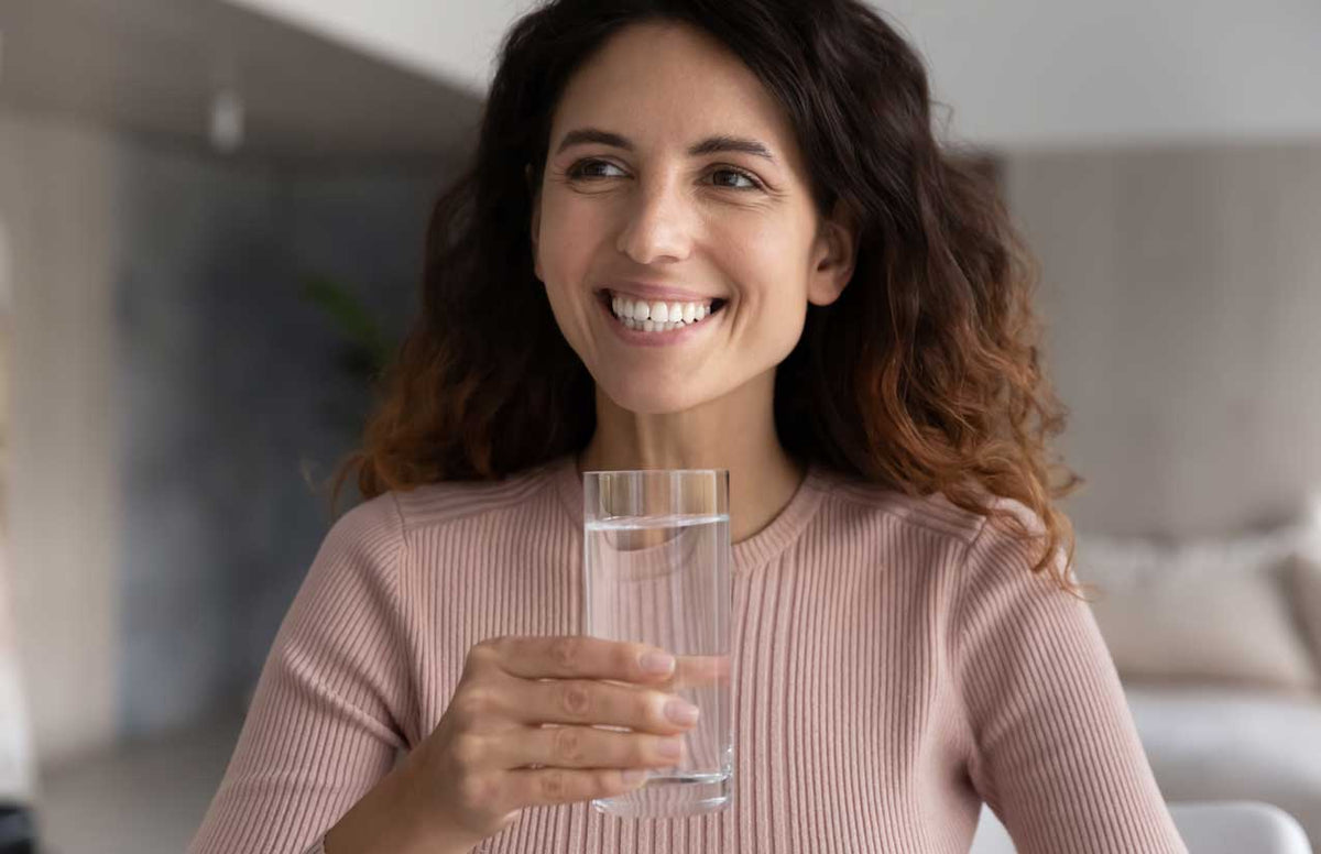 Lächelnde Frau hält im Wohnraum ein Glas mit klarem Wasser in der Hand