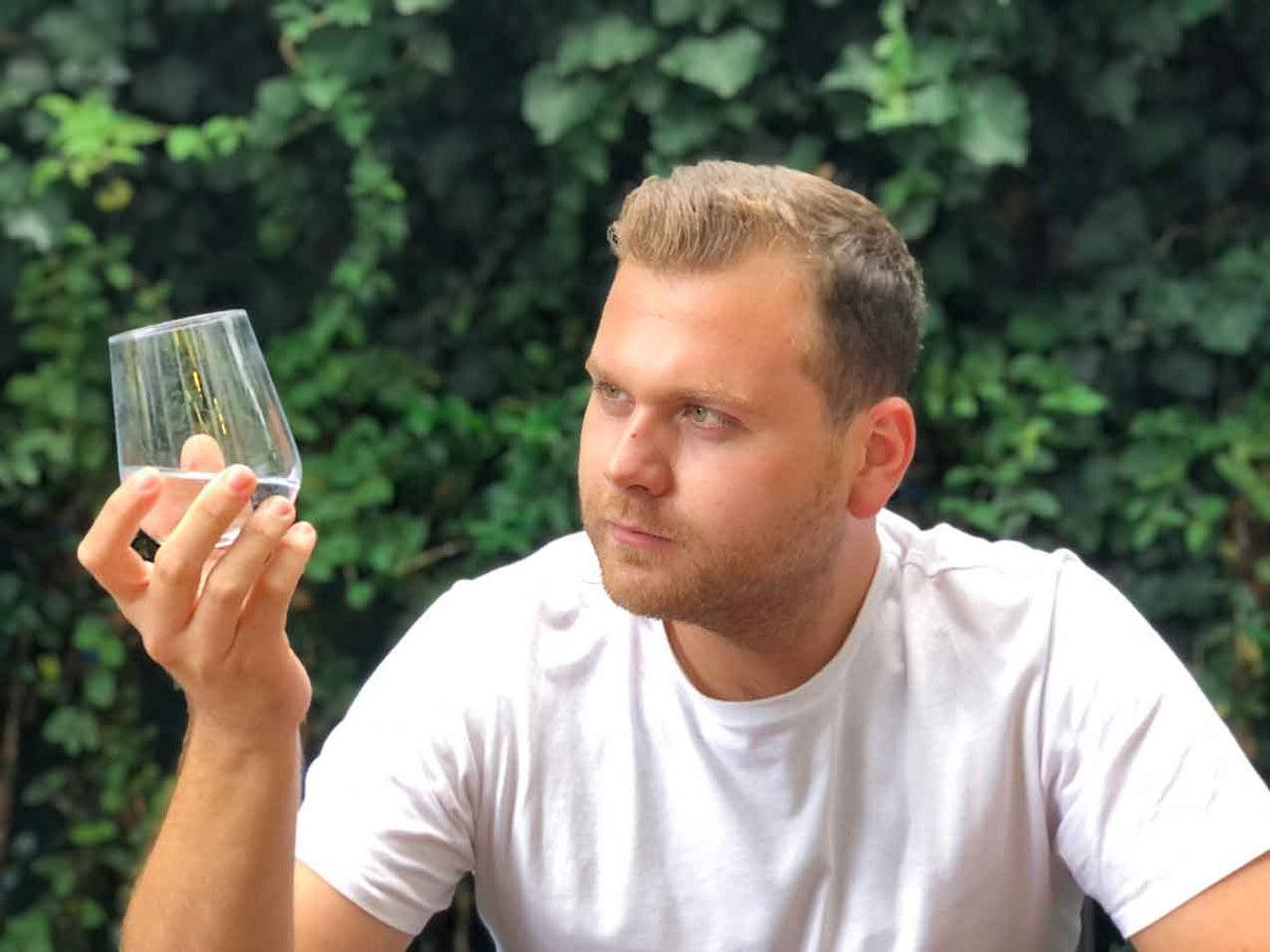 Mann im weißen Shirt hält ein Wasserglas gegen das Licht und prüft dessen Färbung