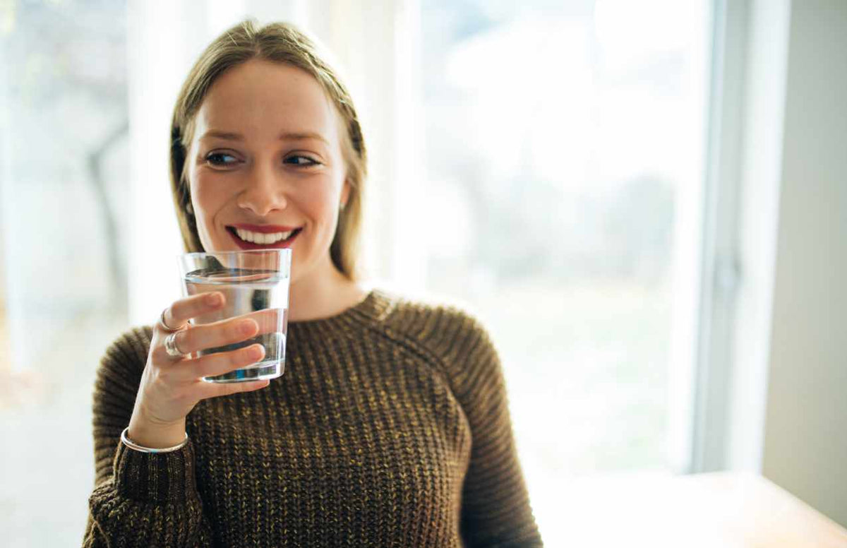 Lächelnde junge Frau am Fenster hält ein Glas klares Trinkwasser in der Hand