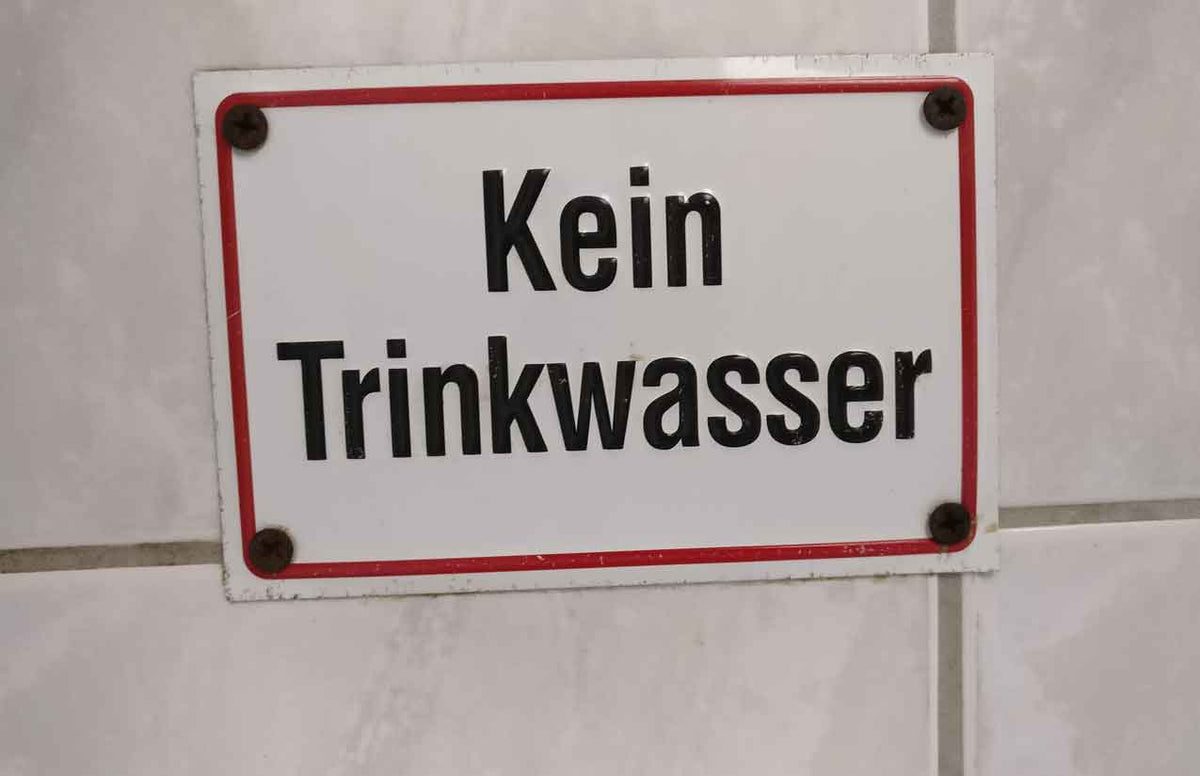 Weißes Emailleschild mit rotem Rand und der Aufschrift „Kein Trinkwasser“ an einer gefliesten Wand