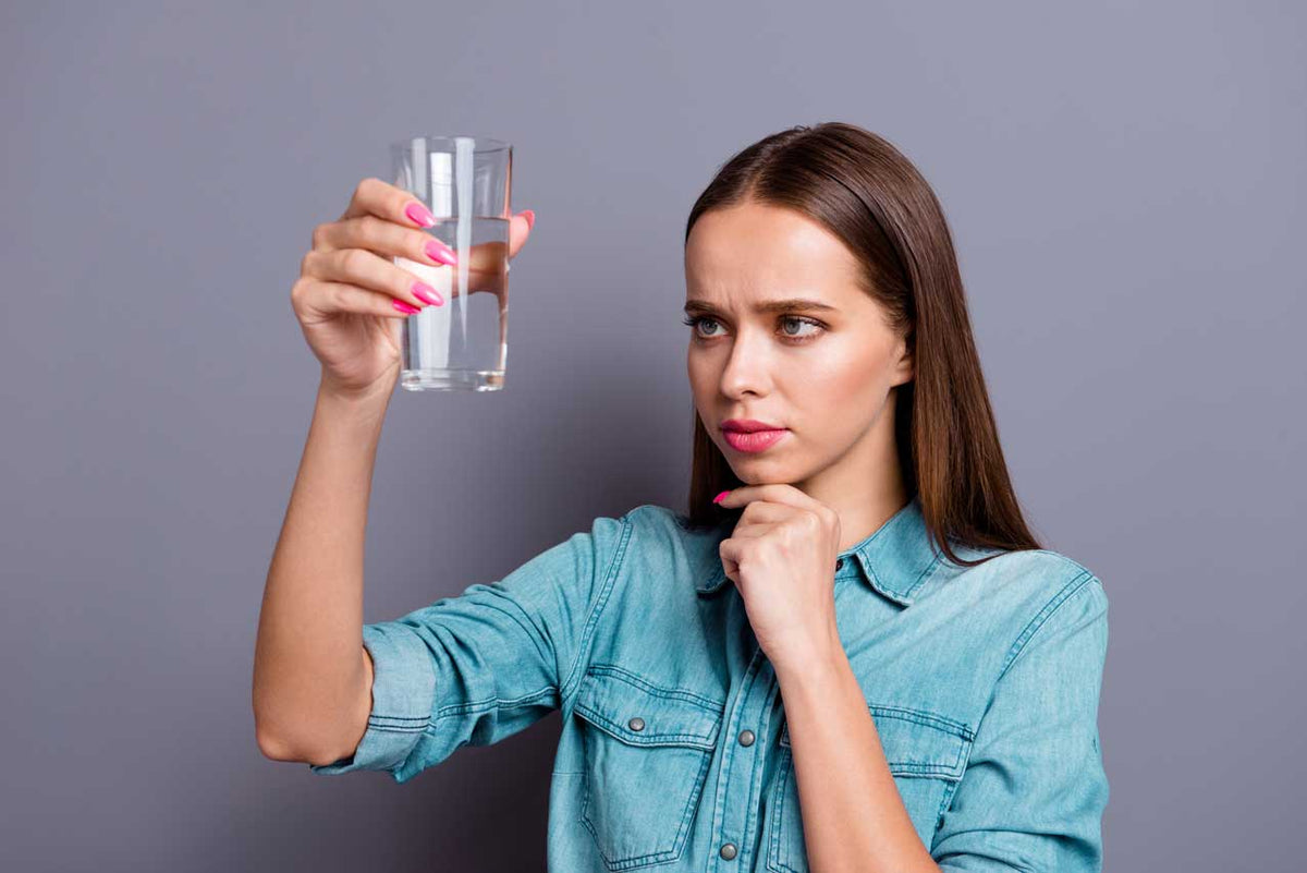 Frau im Jeanshemd hält ein Glas Wasser hoch und mustert es zweifelnd, Hand am Kinn