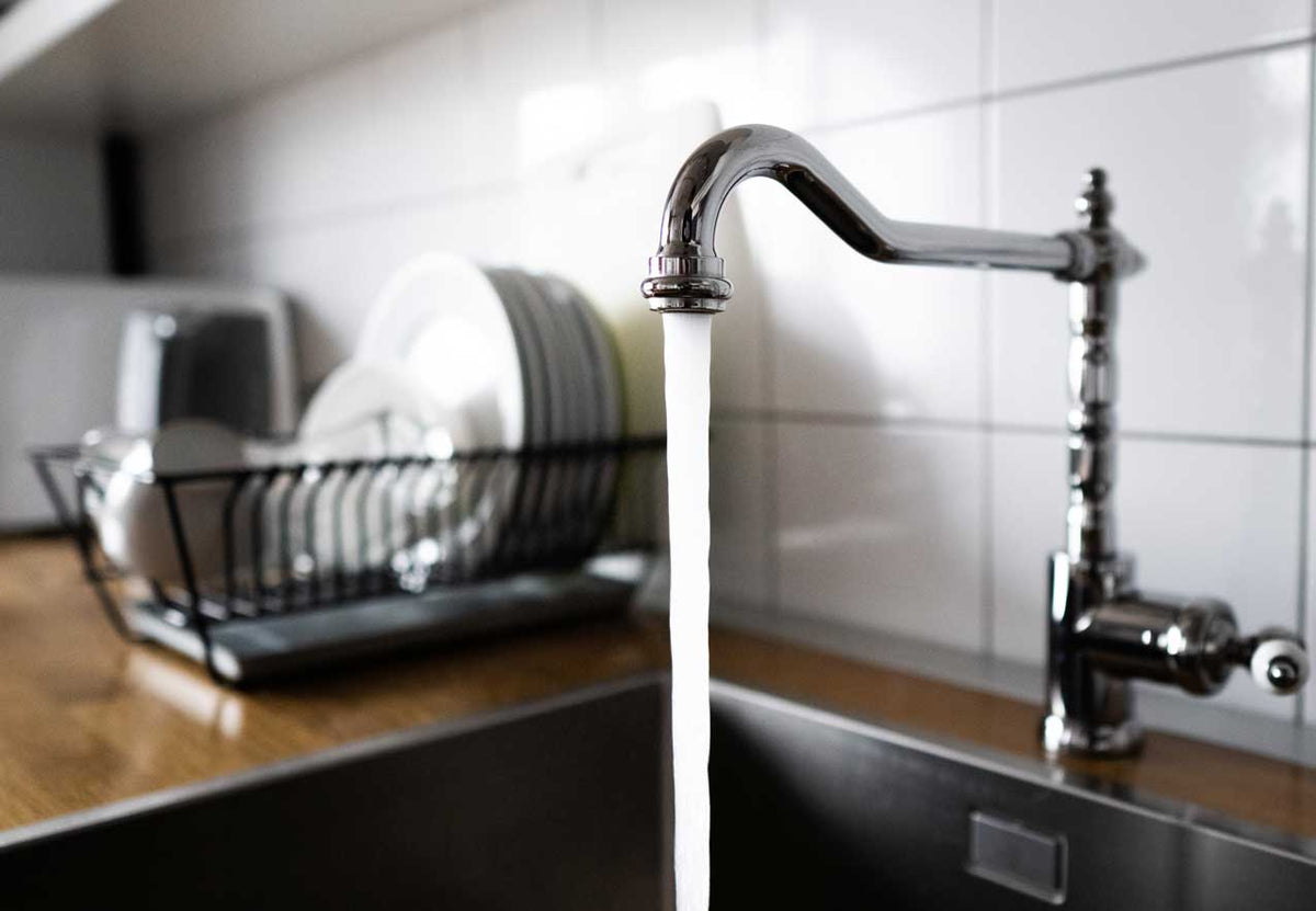 Wasserstrahl läuft aus einer Küchenarmatur in die Spüle, Geschirrständer im Hintergrund