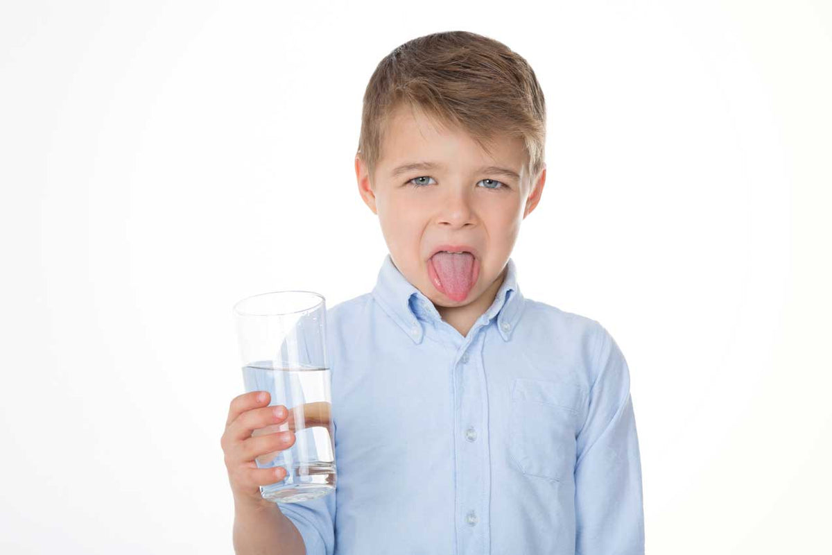 Junge im hellblauen Hemd streckt angewidert die Zunge heraus und hält ein Glas Wasser