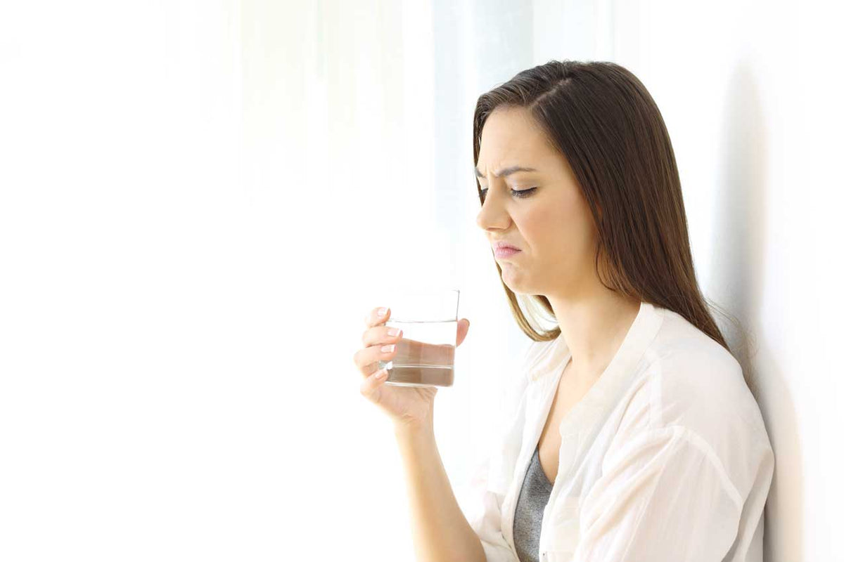 Frau verzieht den Mund und hält ein Glas Wasser vor einer hellen Wand