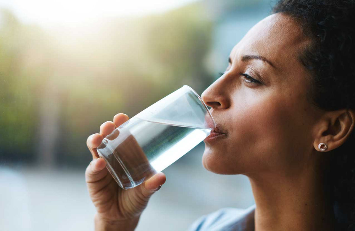 Frau trinkt im Profil draußen ein Glas klares Wasser, grüner Hintergrund unscharf