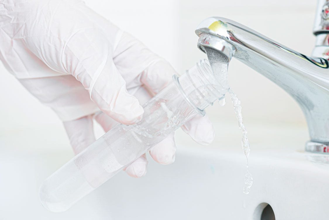Behandschuhte Hand füllt ein Probenröhrchen mit Wasser aus dem Wasserhahn am Waschbecken