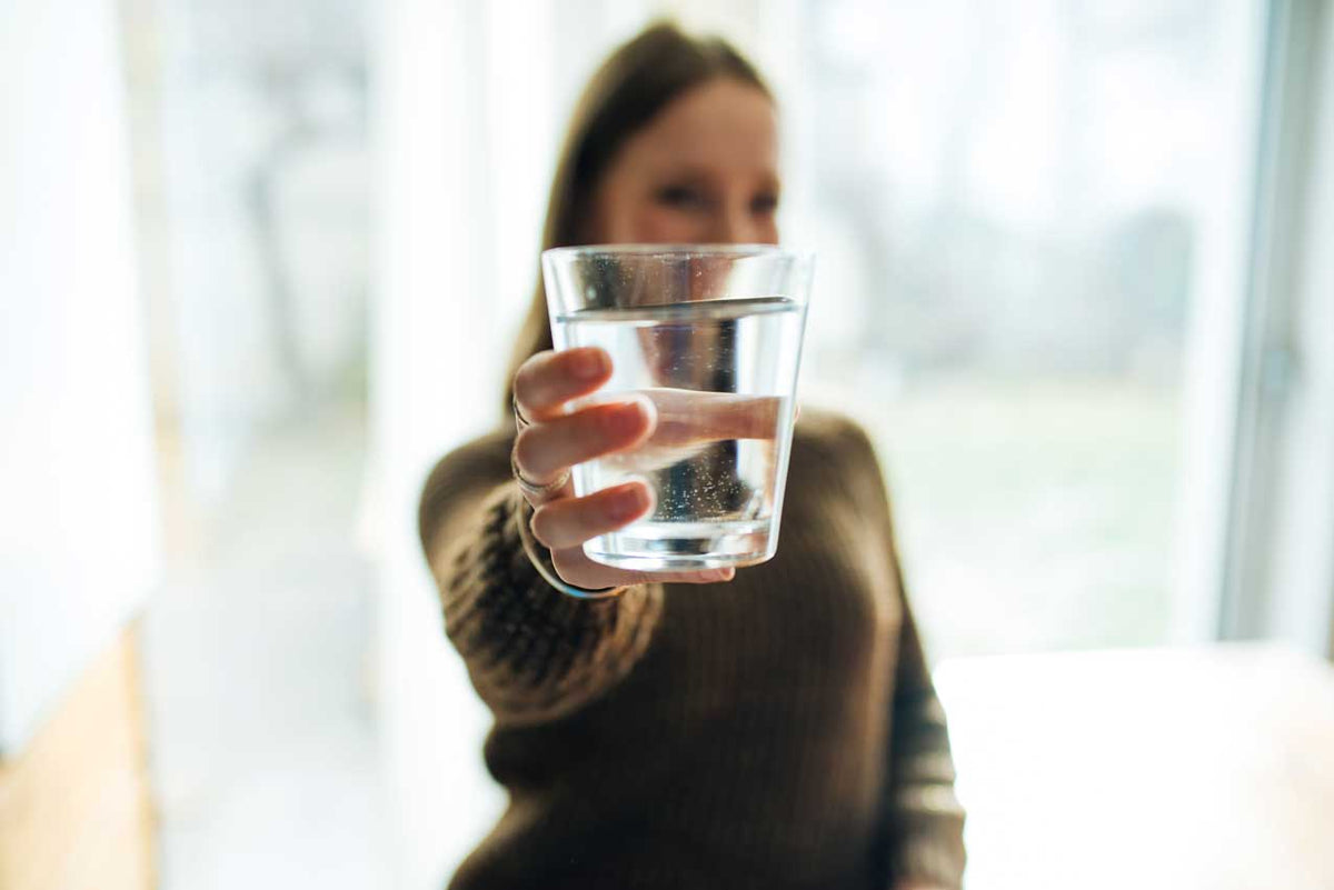Frau streckt am Fenster ein gefülltes Wasserglas in Richtung Kamera, Hintergrund unscharf