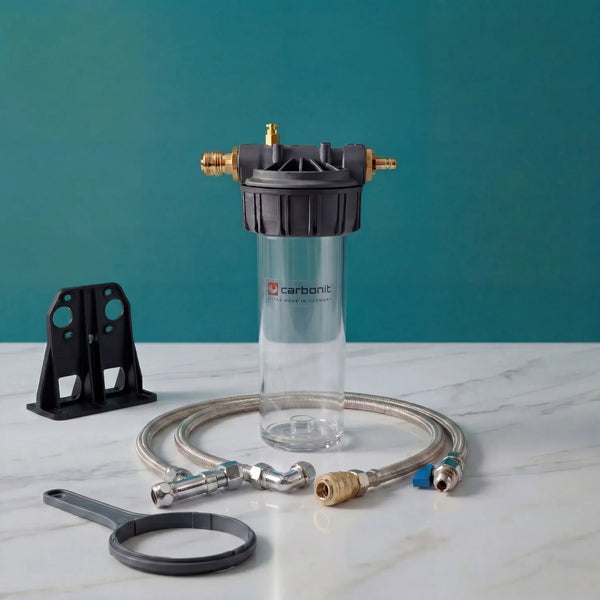 Carbonit Vario Basic Einbaufilter-Set auf weißer Marmorarbeitsplatte – transparentes Filtergehäuse (leer) mit schwarzem Kunststoffkopf und zwei Messing-Schnellkupplungen, Carbonit-Logo auf Gehäuse, schwarzer Wandhalterung, grauer Ringschlüssel, Schlauch mit Winkelverbinder und Kugelventil; teal Hintergrund
