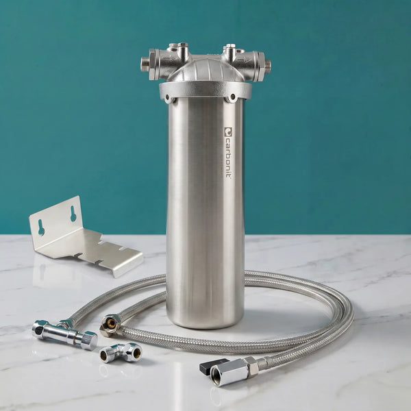 Carbonit Vario Inox VA Einbaufilter-Set auf weißer Marmorarbeitsplatte – gebürstetes Edelstahlgehäuse mit Carbonit-Logo, Gusskopf mit zwei Gewindeanschlüssen, Edelstahl-Wandhalter (L-Form), langer gewickelter Edelstahlschlauch, Winkelstücke und Fittings; teal Hintergrund