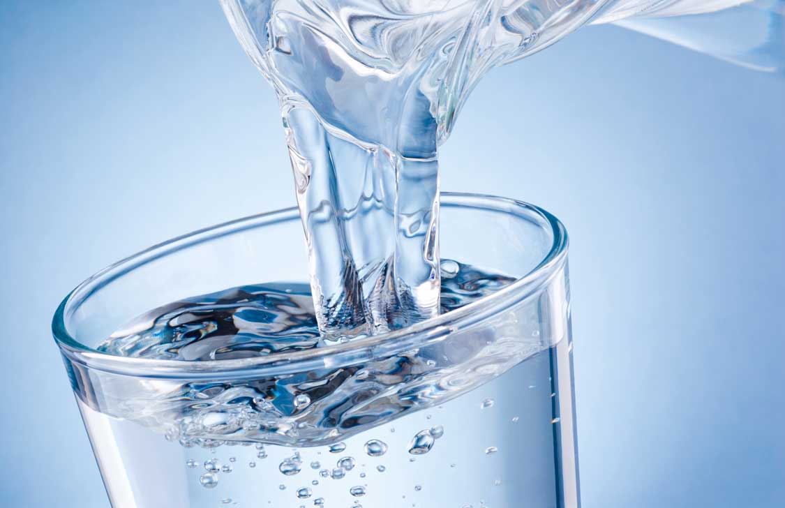 Wasser wird aus einer Glaskaraffe in ein randvolles Trinkglas gegossen