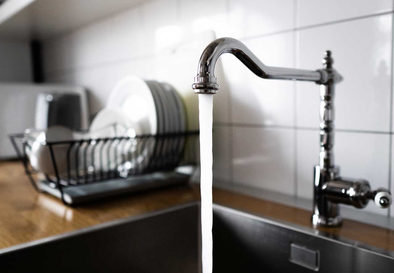 Wasserstrahl läuft aus einer Küchenarmatur in die Spüle, Geschirrständer im Hintergrund