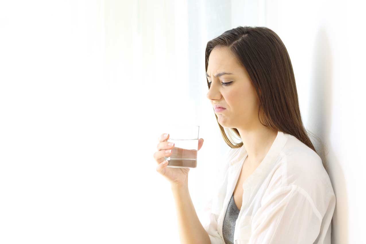Frau verzieht den Mund und hält ein Glas Wasser vor einer hellen Wand