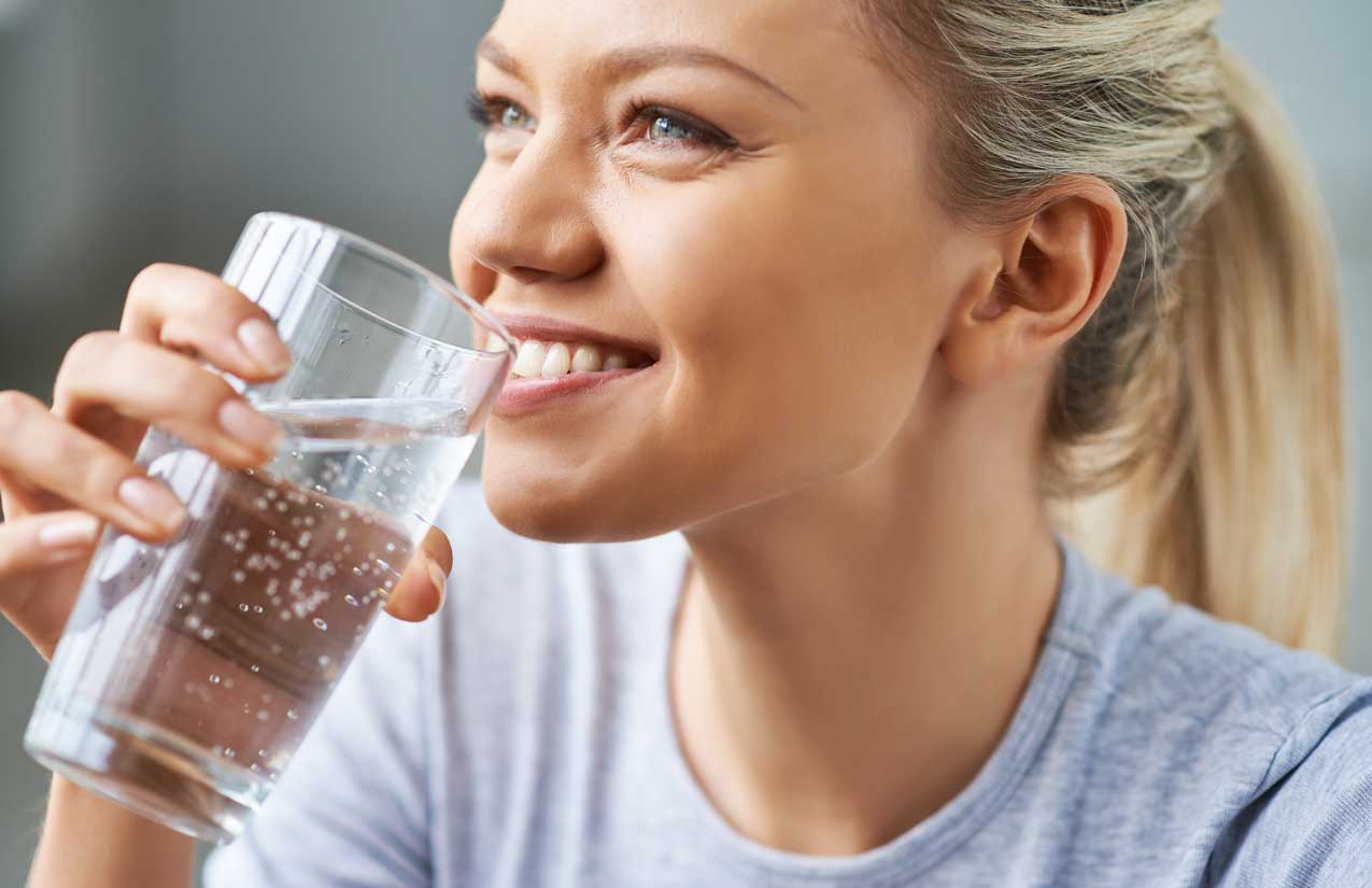 Junge Frau trinkt lächelnd aus einem Glas mit perlendem Wasser, Nahaufnahme