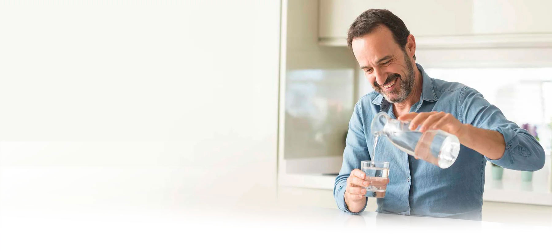 Lächelnder Mann gießt frisches Wasser aus einem Glaskrug in ein Wasserglas – gesundes Trinken mit Vitalhelden