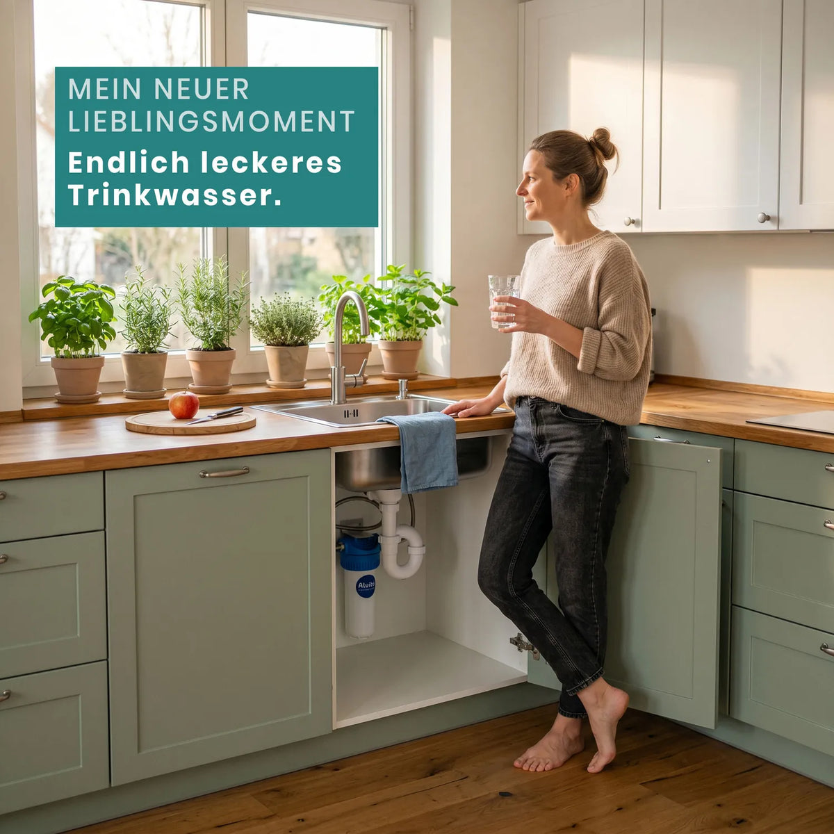 Frau steht barfuß in Küche mit grünen Holzschränken und Holzarbeitsplatte, hält Glas gefiltertes Wasser; Kräutertöpfe am Fenster; offener Unterschrank zeigt installierten Smart Direct Einbaufilter; Text-Overlay: „Mein neuer Lieblingsmoment – Endlich leckeres Trinkwasser."