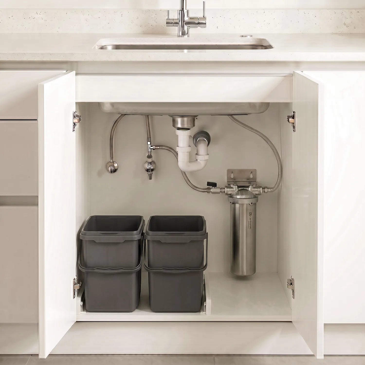 Carbonit Vario Inox VA im offenen Unterschrank – gebürstetes Edelstahlgehäuse mit Gusskopf an weißer Kücheninnenwand montiert; Schlauchverbindungen und Absperrventile sichtbar; weiße Küche mit Kreuzgriff-Armatur und Marmorarbeitsplatte