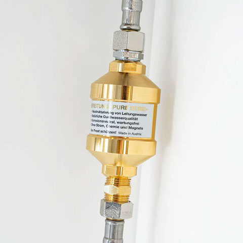 Goldener UMH Vitalisierer der Pure Serie zwischen zwei Edelstahlschläuchen an weißer Wand montiert